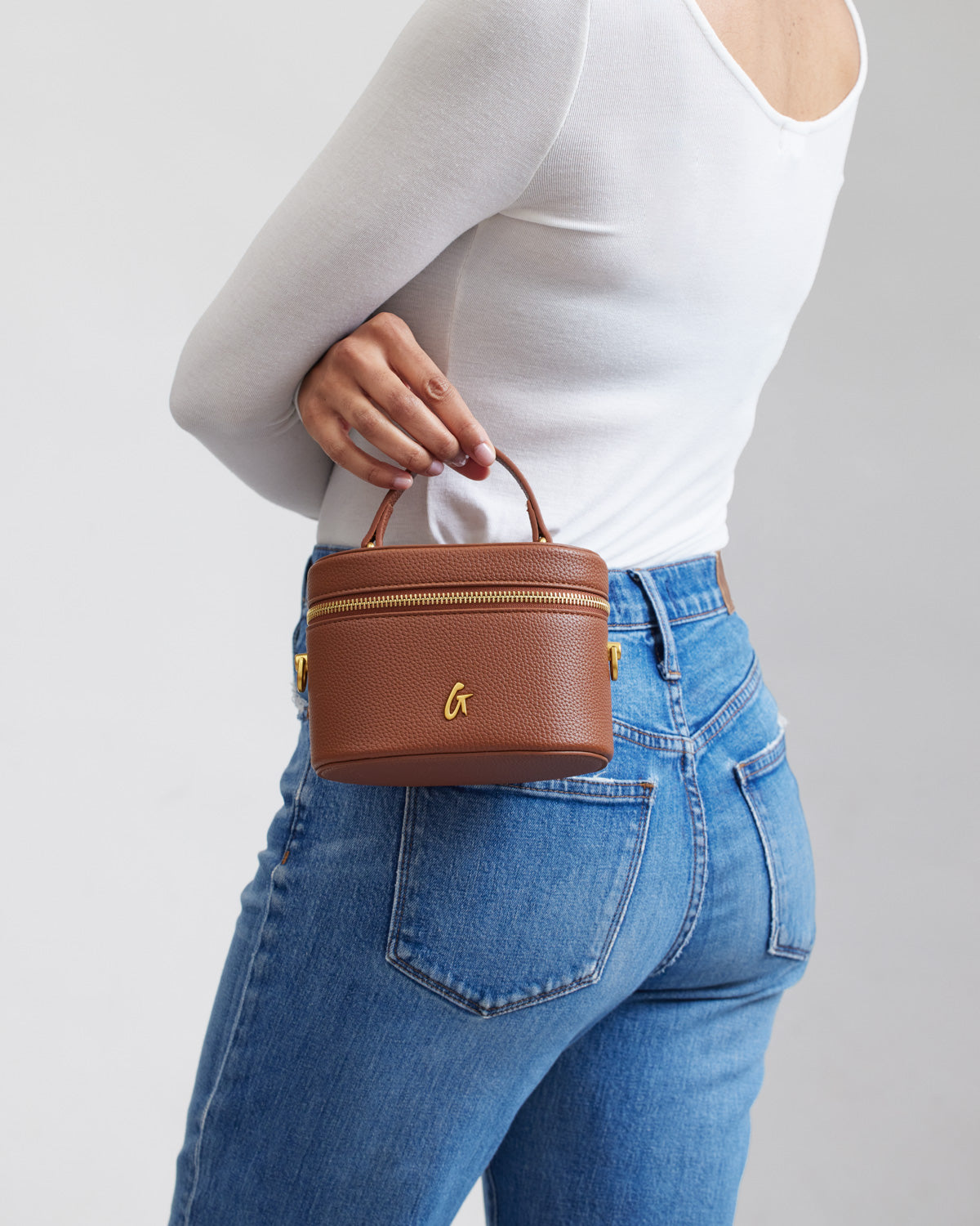 Mini pebble brown vanity bag with gold zipper and logo, held by a woman in a white top and blue jeans.