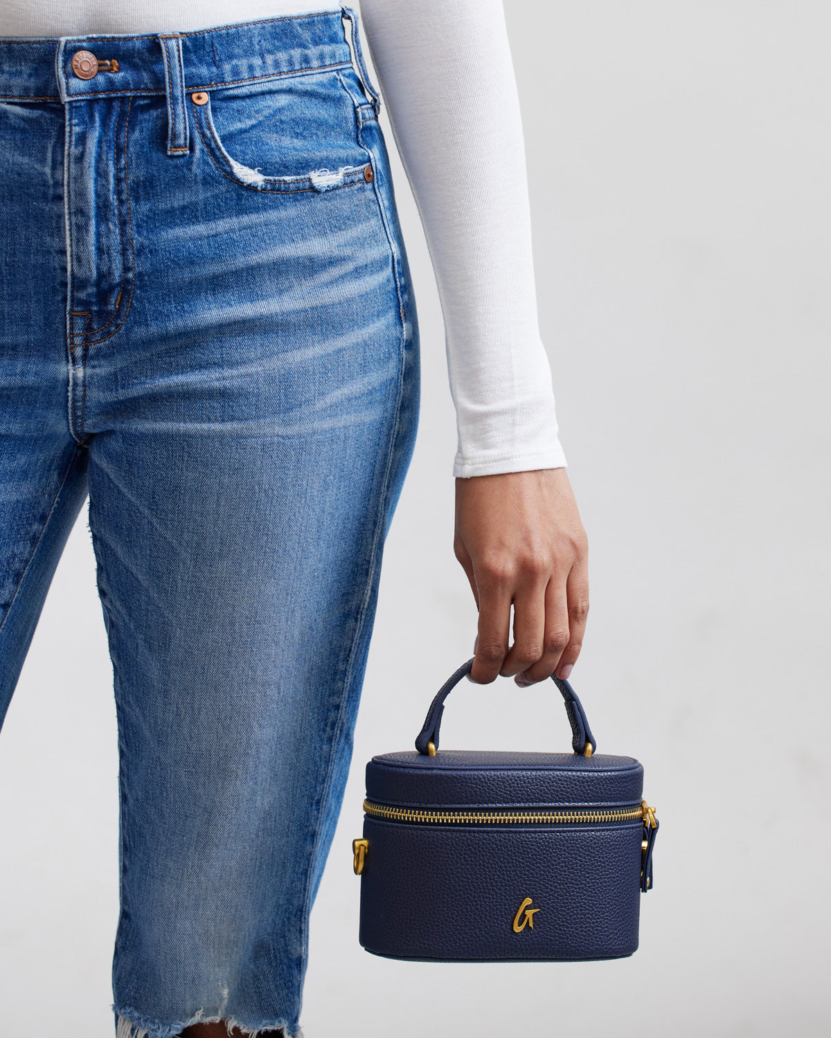 Mini navy pebble vanity bag with gold zipper and handle, held by a person wearing blue jeans and a white long-sleeve shirt.