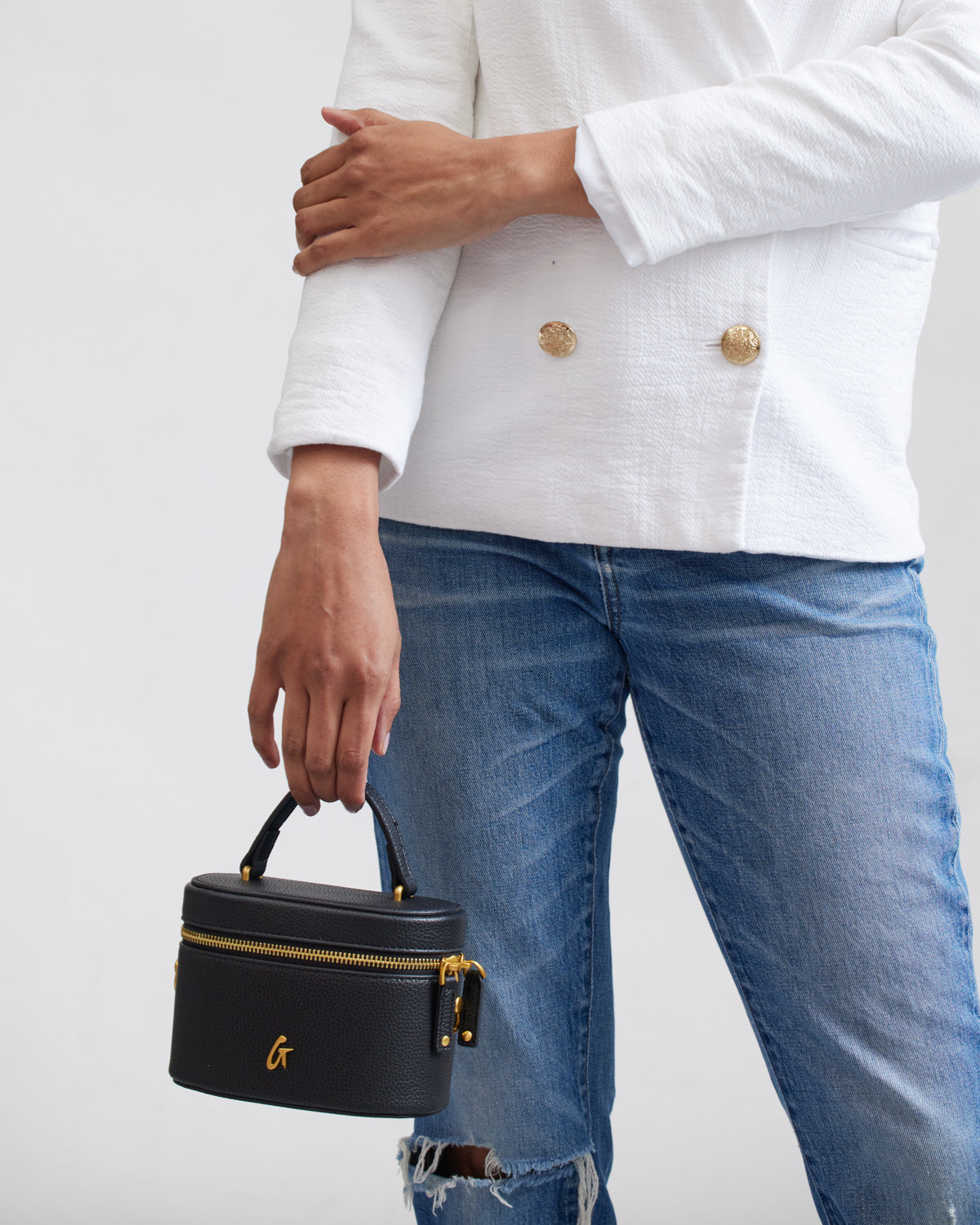 Small black mini pebble vanity bag with gold zipper and handle, held by a person wearing a white blazer and ripped jeans.