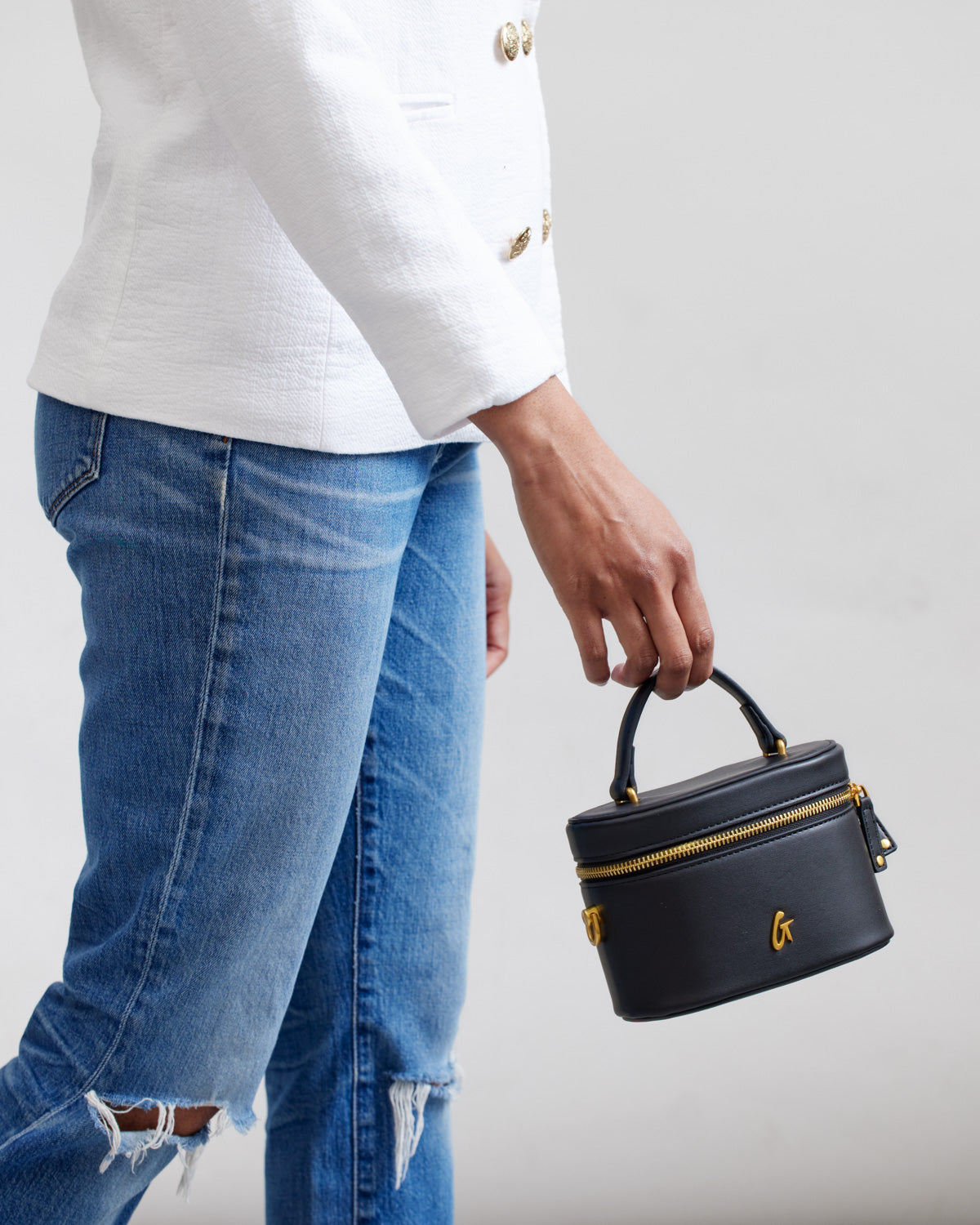 Mini black vanity bag with gold accents, top handle, and zipper closure, held by a person wearing a white blazer and ripped jeans.