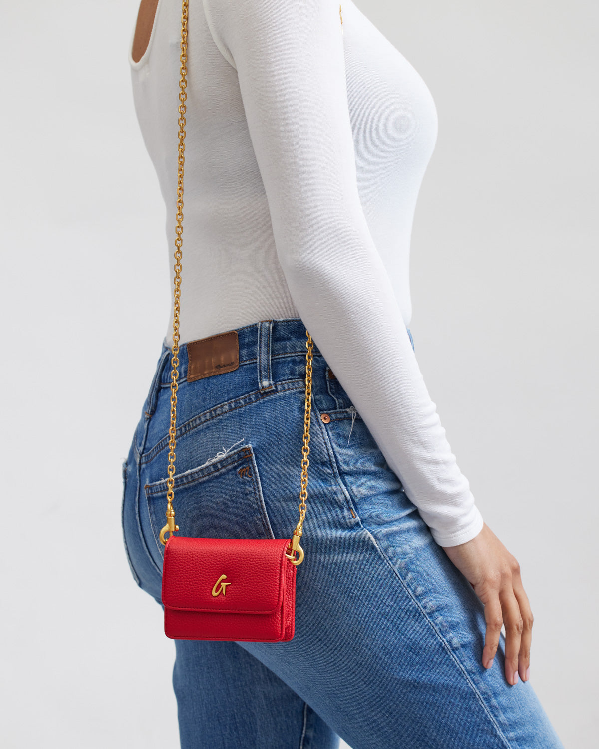 Red MINI PEBBLE WOC crossbody bag with a gold chain strap and gold "G" logo, worn over a white long-sleeve top and blue jeans.