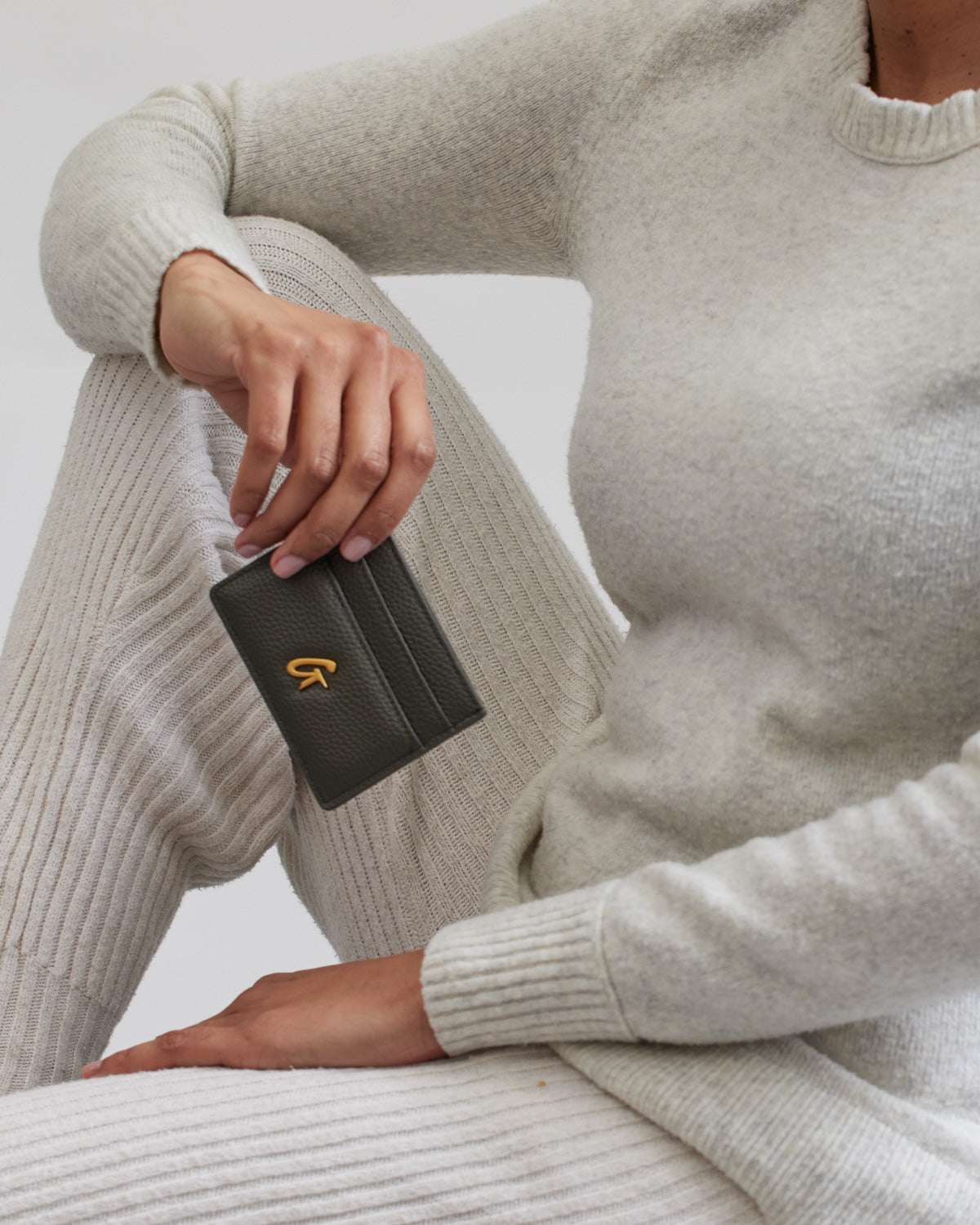 Olive green pebble card holder with a gold logo, held by a person dressed in a cream-colored knit outfit.