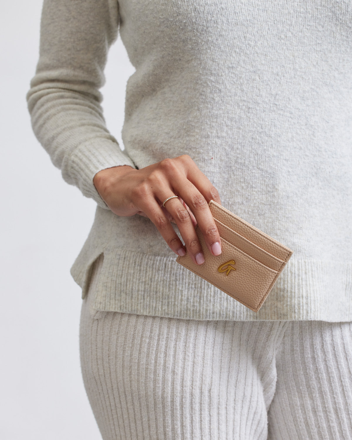 A person holding a nude pebble leather card holder with a gold logo, dressed in a beige sweater and matching pants.