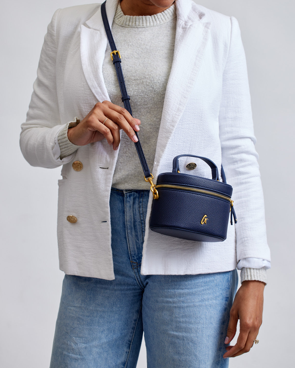 Mini navy pebble vanity bag with gold zipper and logo, worn crossbody by a person in a white blazer and jeans.