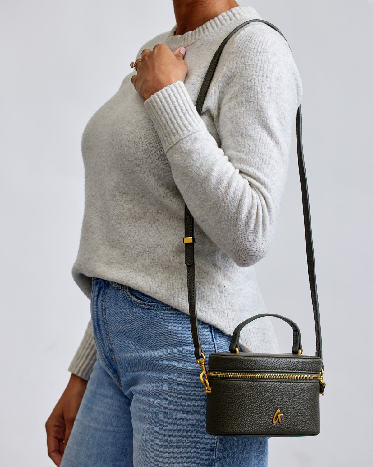 Mini pebble olive green vanity bag with gold accents, carried by a woman in a light sweater and jeans.