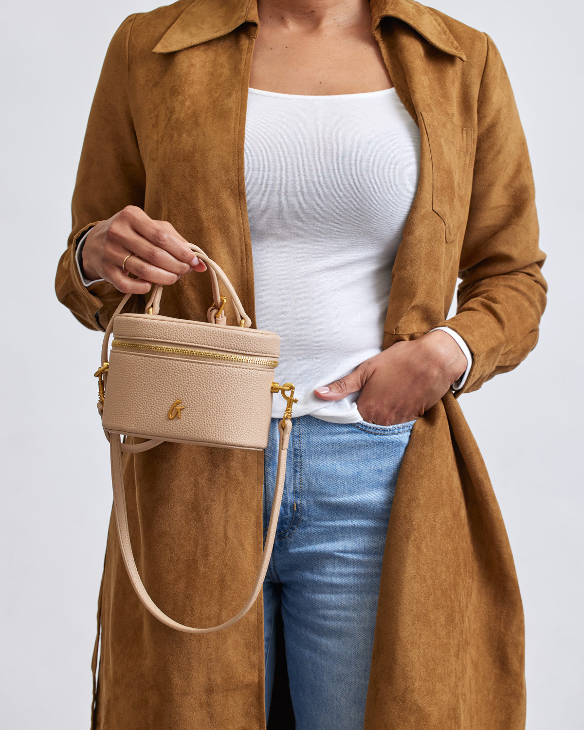 Mini pebble nude vanity bag with gold zipper and logo, held by a woman in a white top and brown coat.
