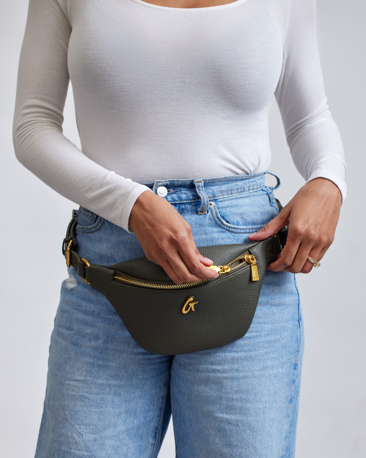 Mini pebble olive green fanny pack with gold zipper and logo, worn around the waist over blue jeans and a white long-sleeve top.