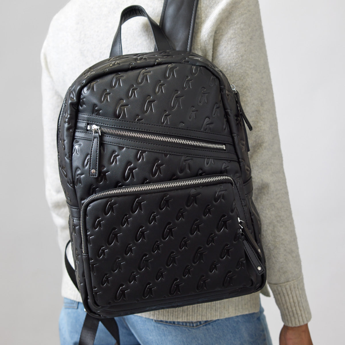 Black unisex backpack with embossed monogram pattern, multiple zippered compartments, and adjustable straps.