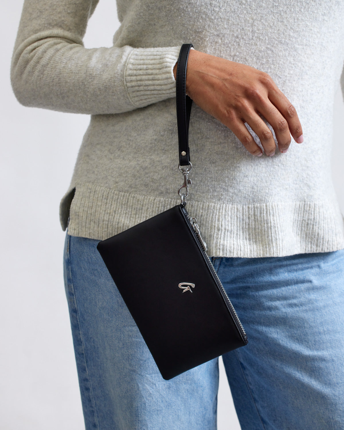 Mini silver wristlet in black with a silver zipper and logo, attached to a black strap, worn on a person's wrist.