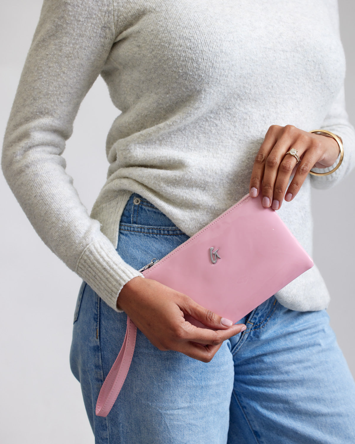 Mini silver wristlet with mirror pink finish, featuring a silver logo, held by a person in a beige sweater and blue jeans.