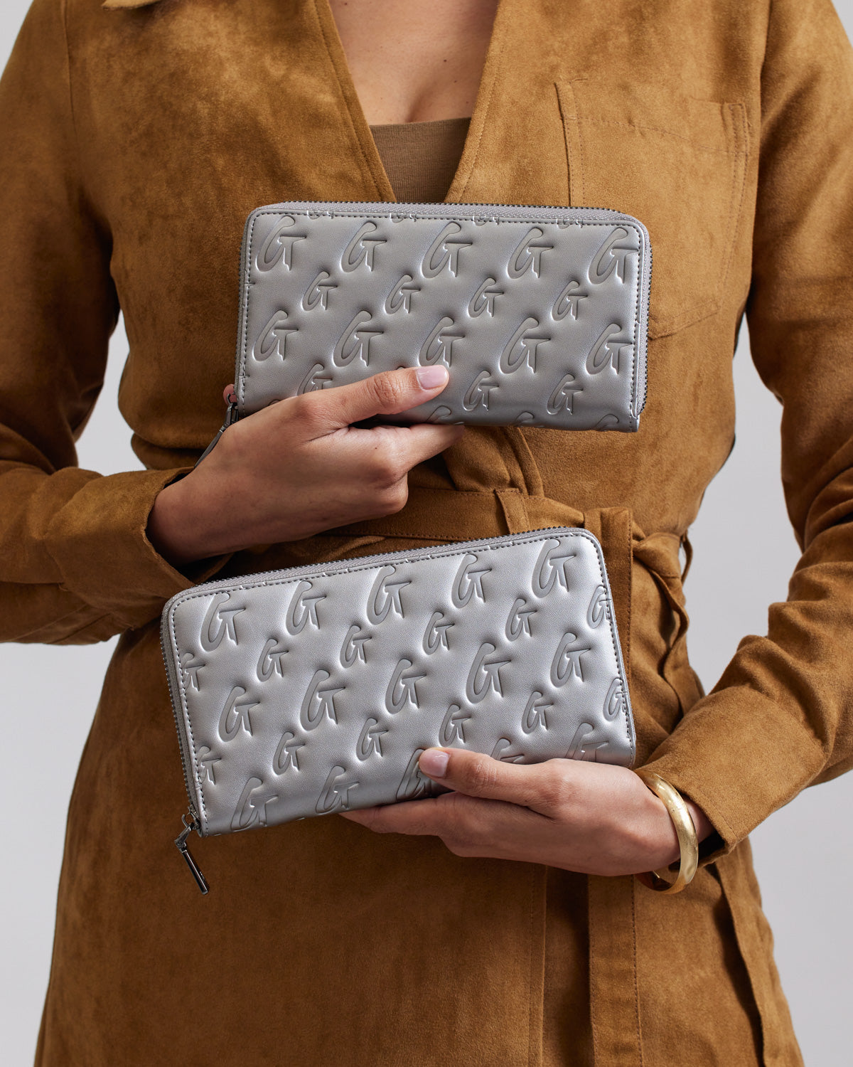 Two silver monogram wallets with embossed G patterns, held by a person wearing a brown suede coat.