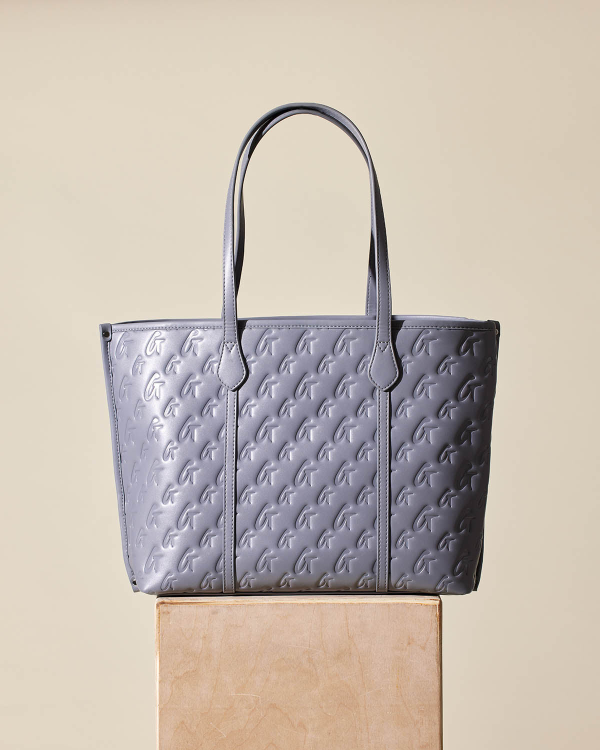 Gray Monogram Sleek Tote bag with embossed logo pattern, dual handles, and structured design, displayed on a wooden block.