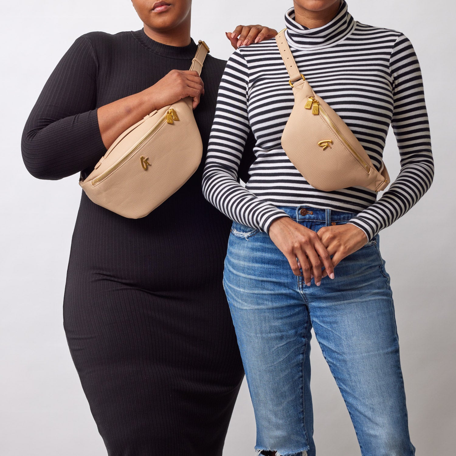 Two women wear beige Pebble Fanny Packs with gold zippers and logo, styled over casual black and striped outfits.