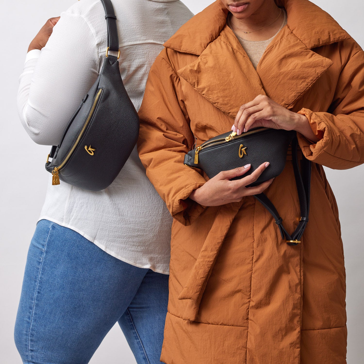 Two women display black Pebble Fanny Pack Bundles with gold accents, one worn crossbody and the other held in hands.