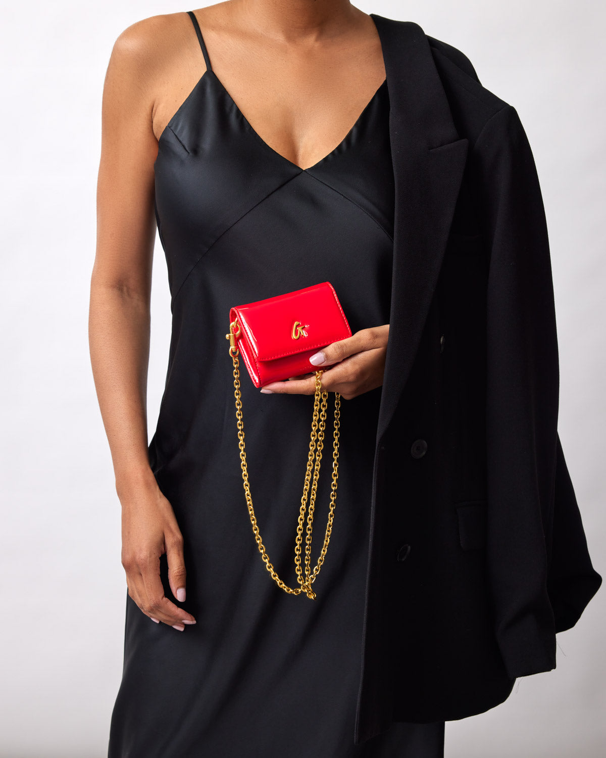 Red compact mirror with gold chain and logo, held by a woman in a black satin dress and blazer.