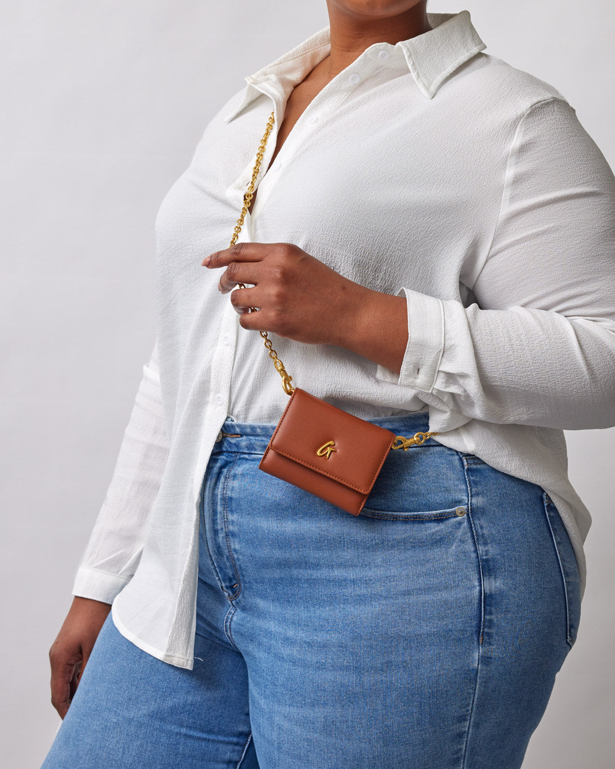 Brown compact wallet with gold logo, attached to a gold chain, worn as a crossbody accessory by a person in casual attire.