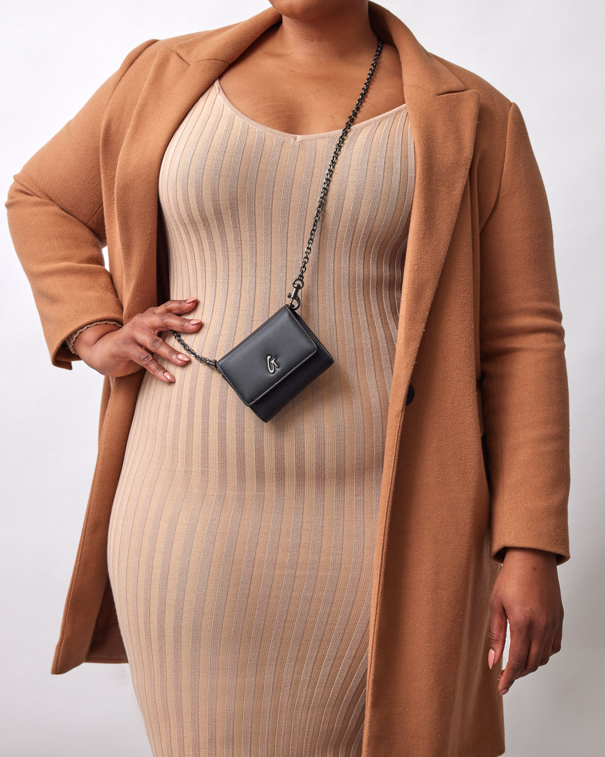 A woman wearing a beige striped dress and camel coat, holding a black compact WOC bag with gunmetal hardware on a chain strap.