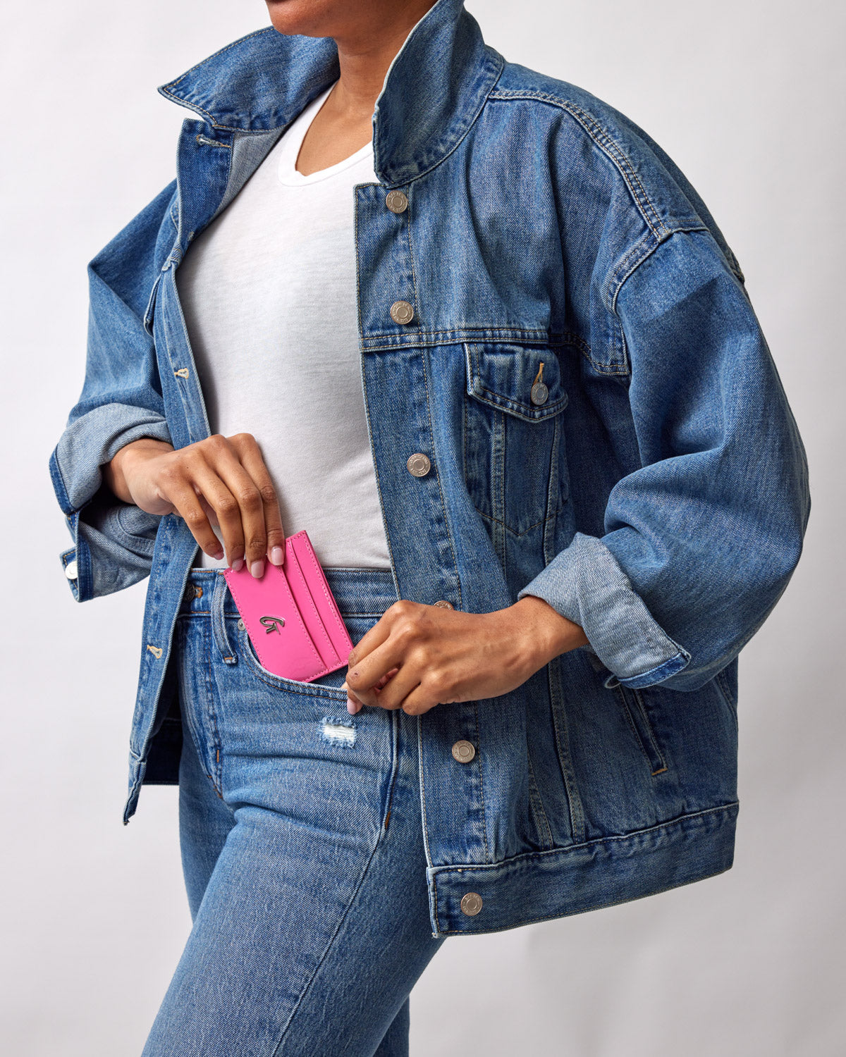 A person in a denim jacket and jeans holds a bright pink mirror hot pink card holder, partially tucked into their front pocket.