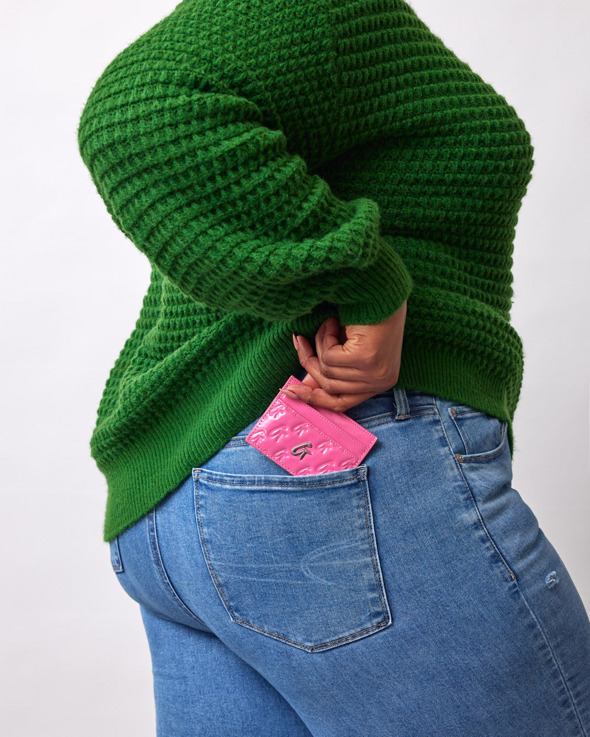A person in a green sweater and blue jeans places a pink mirror hot pink monogram silver card holder into their back pocket.