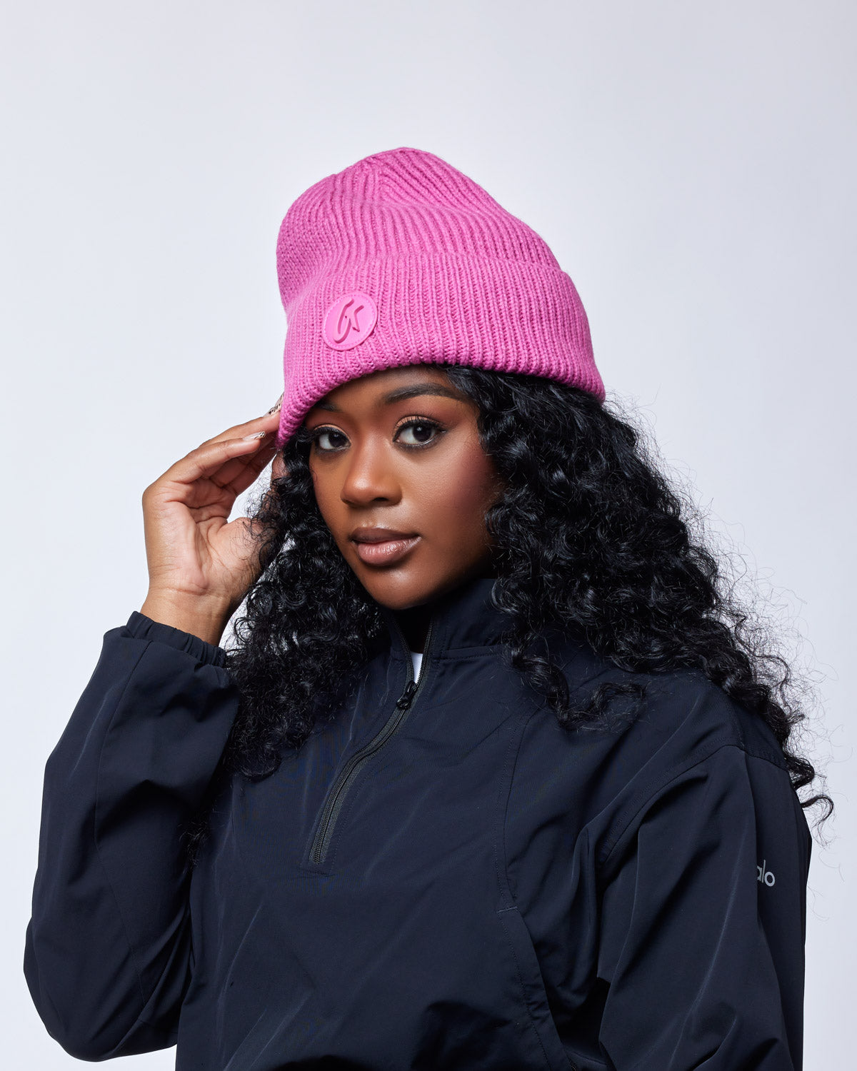 Woman wearing a bright pink classic beanie with a logo patch, dressed in a black jacket, posing against a plain background.