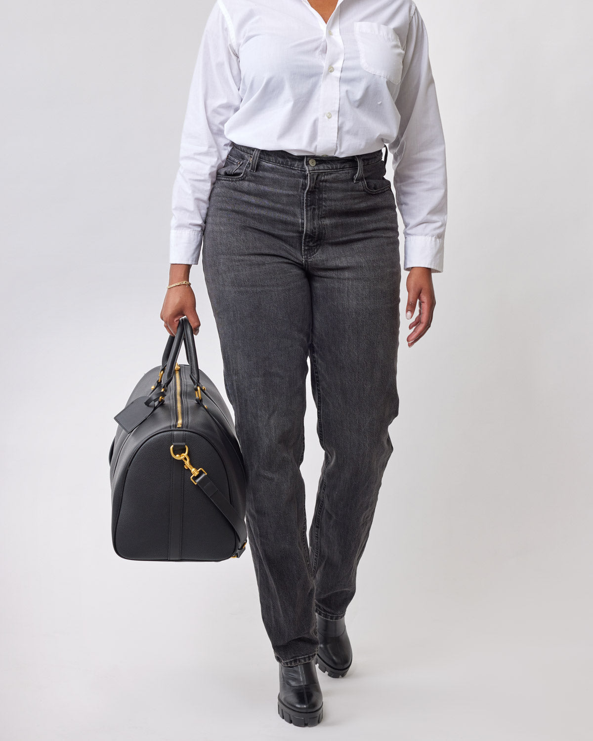 Black Pebble Duffle Bag with gold hardware, held by a person wearing a white shirt and black jeans.