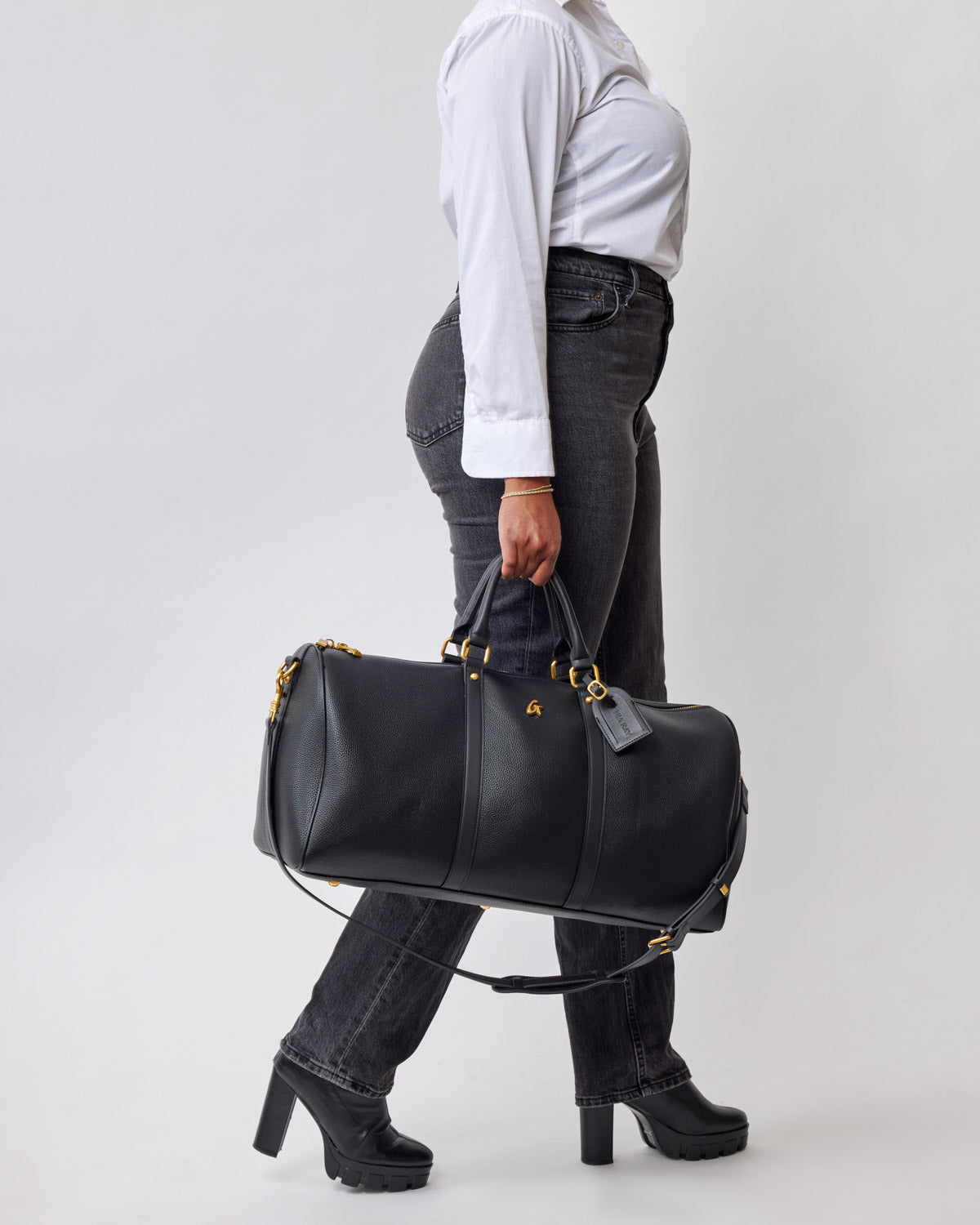 Black Pebble Duffle Bag with gold accents, carried by a person in black jeans, a white shirt, and black heeled boots.