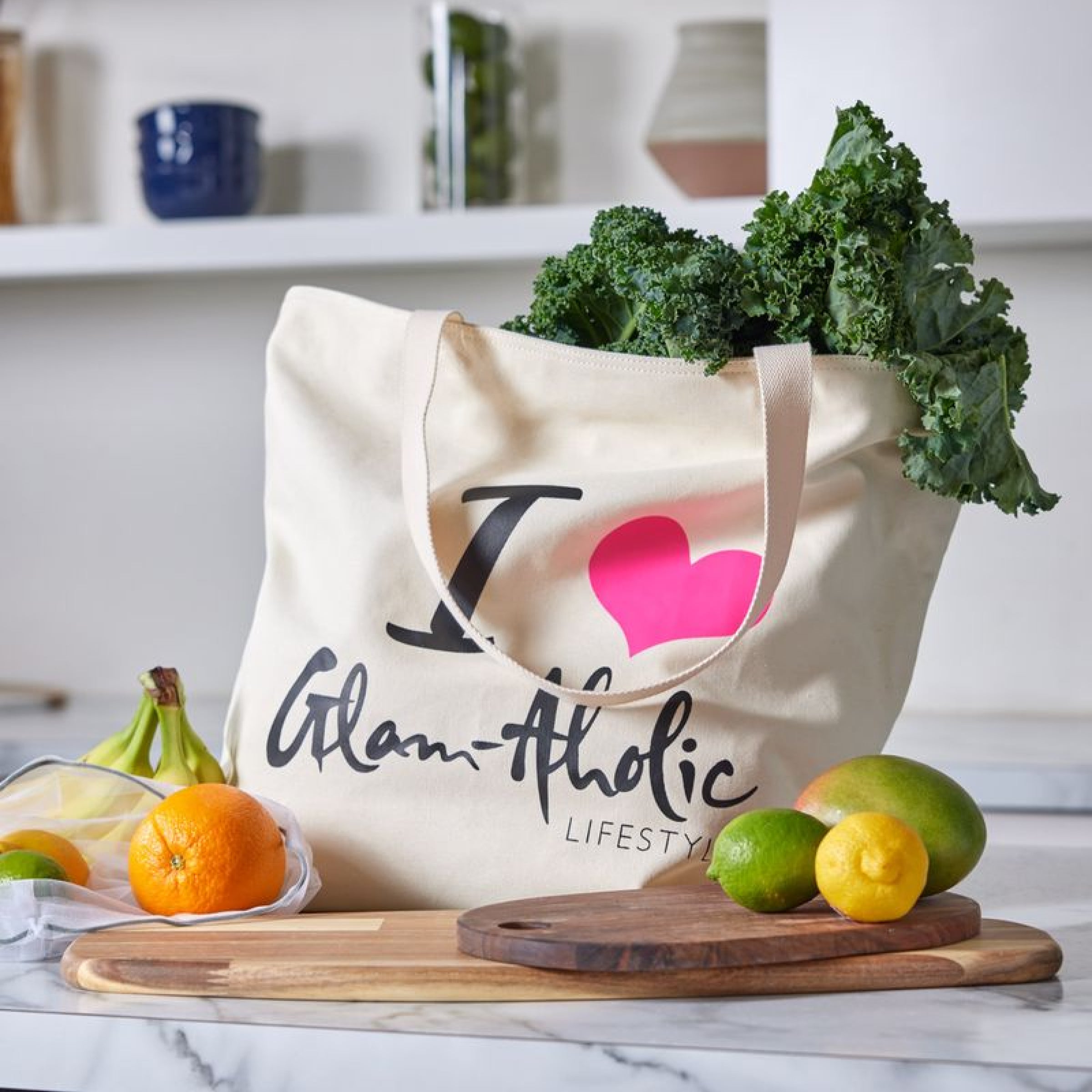Cream tote bag with "I ♥ Glam-Aholic Lifestyle" text, filled with kale, on a wooden cutting board with citrus fruits nearby.