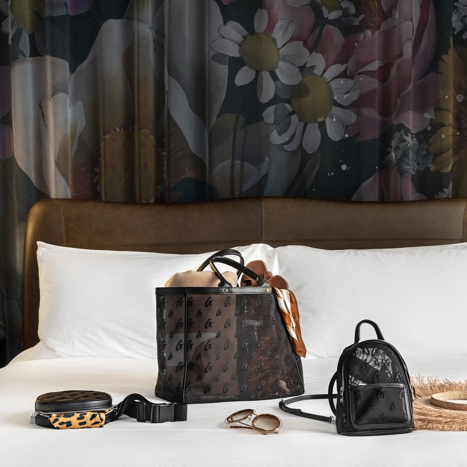 Set of stylish black and leopard print handbags, accessories, and sunglasses on a white bed with floral curtains background.