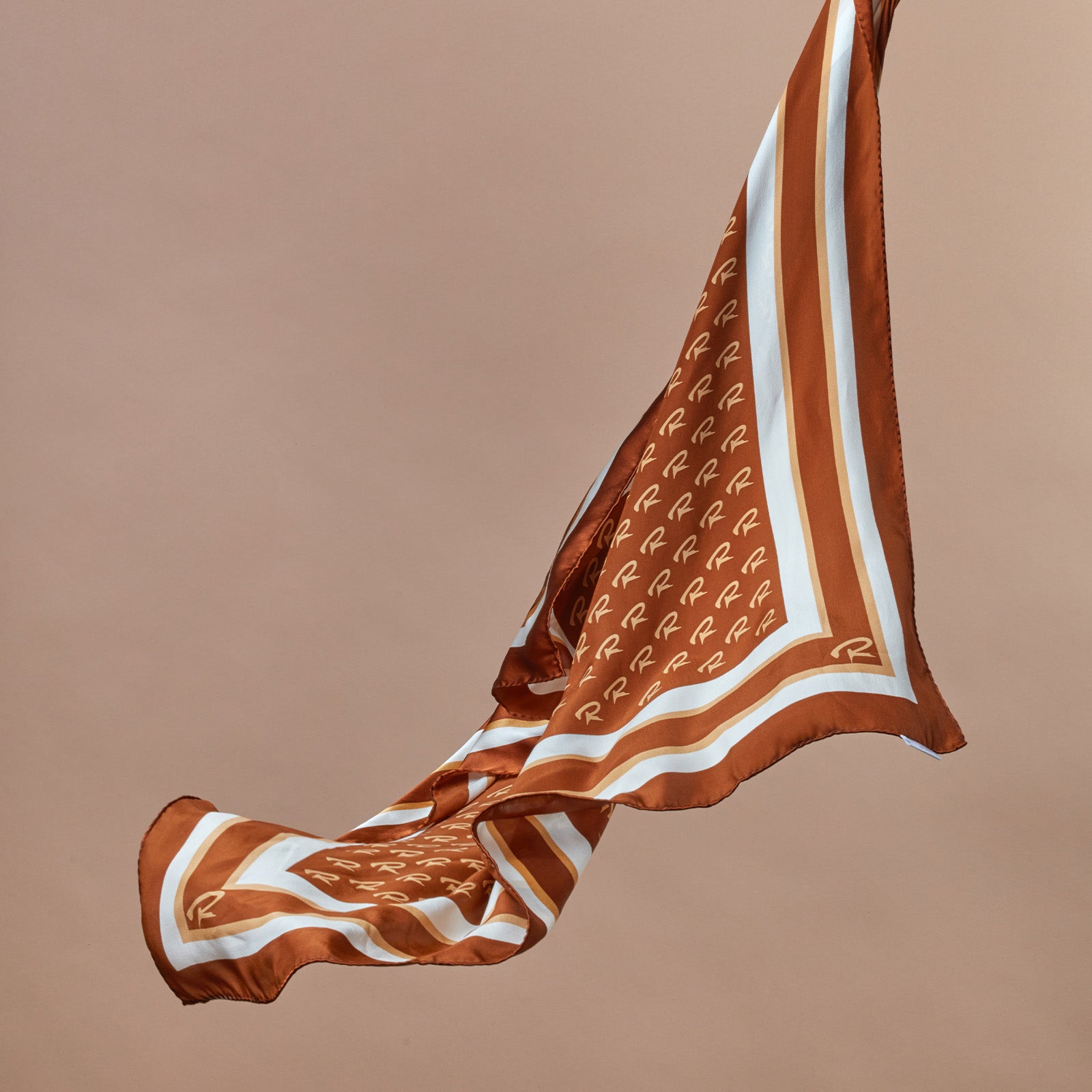 Brown and white patterned silk scarf with "R" monogram and striped border, floating against a beige background.