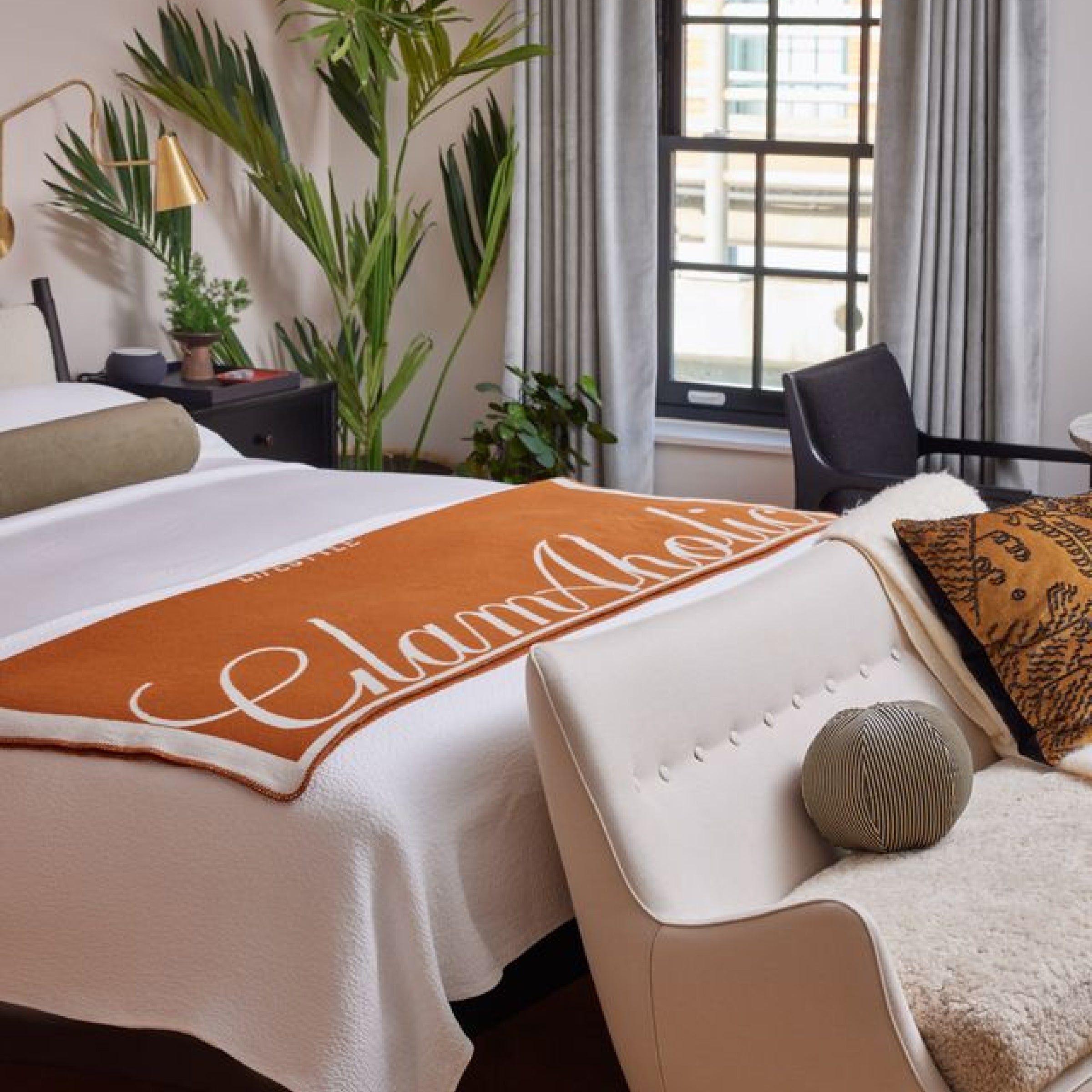 Elegant bedroom with white bed, orange blanket, patterned pillows, black side table, green plants, and large window with c...
