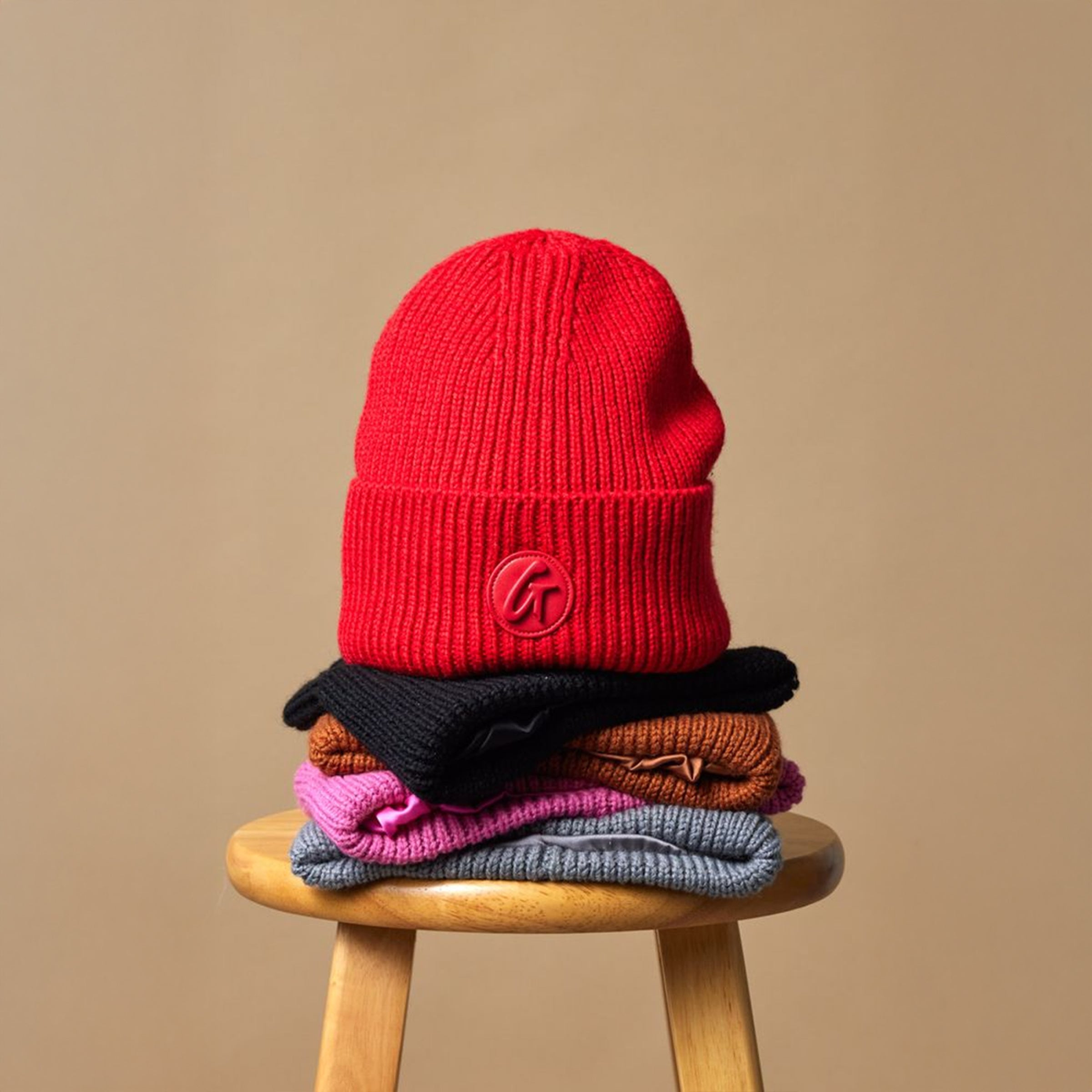 Red knit beanie with a logo patch, stacked on black, brown, pink, and gray sweaters on a wooden stool.
