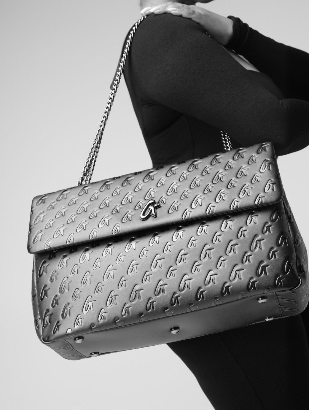 Black quilted handbag with silver chain strap and logo pattern, featuring a front flap and metallic accents.