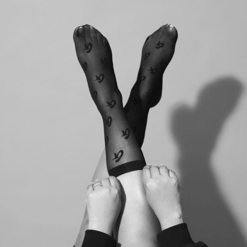 Black sheer stockings with logo print, paired with crossed legs and hands holding calves, creating a stylish, affordable l...