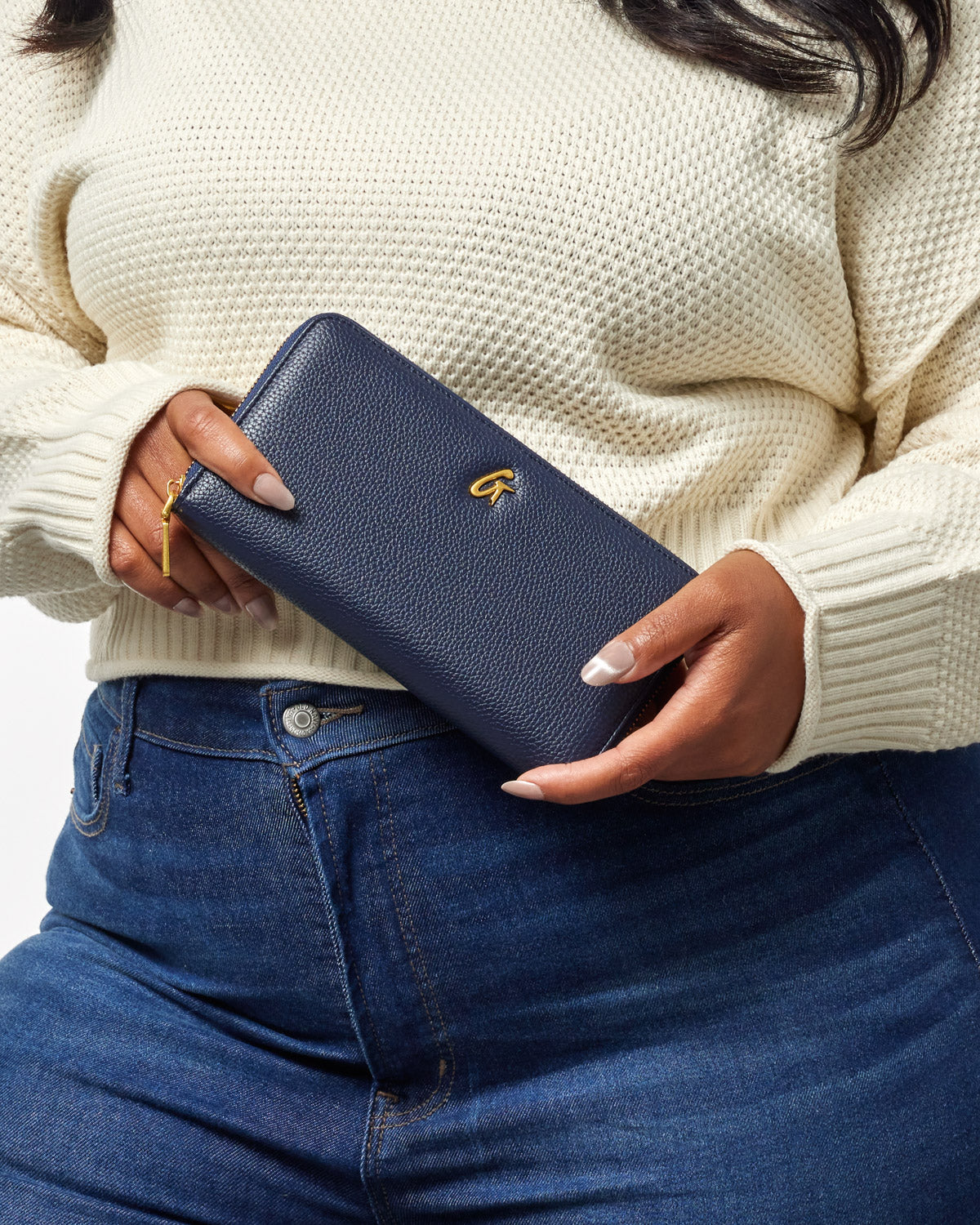 A person holding a navy blue pebble leather wallet with a gold logo, wearing a cream sweater and blue jeans.
