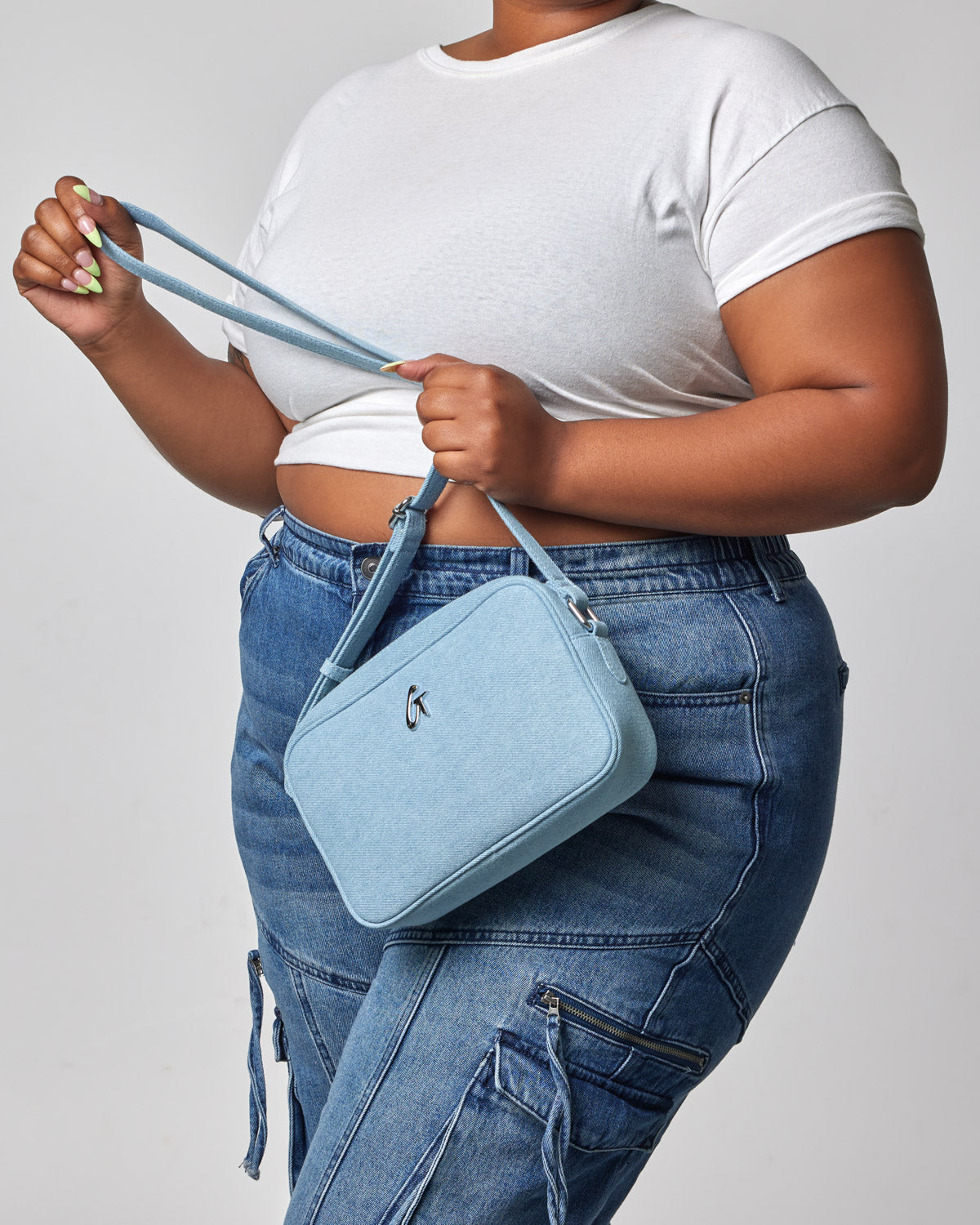 A woman wearing a white t-shirt and jeans holds a light blue denim camera bag with a silver logo, showcasing the product.