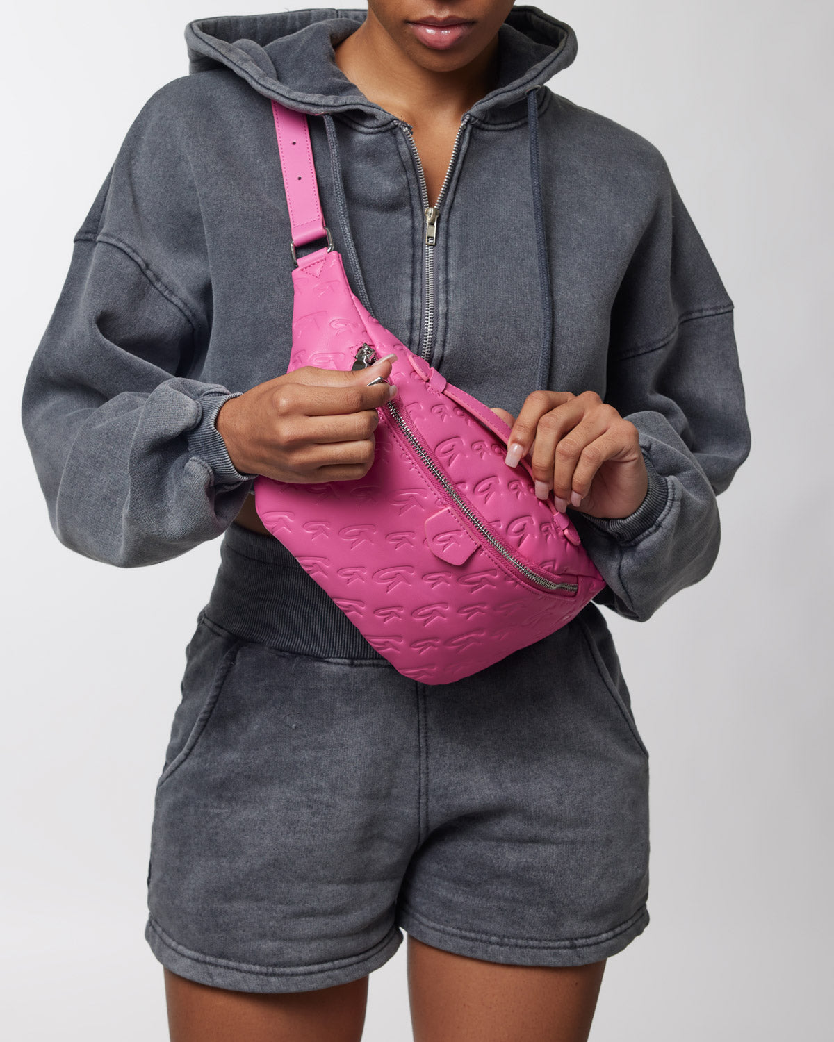 A woman wears a gray hoodie and shorts while holding a bright pink monogram fanny pack with embossed logo details.