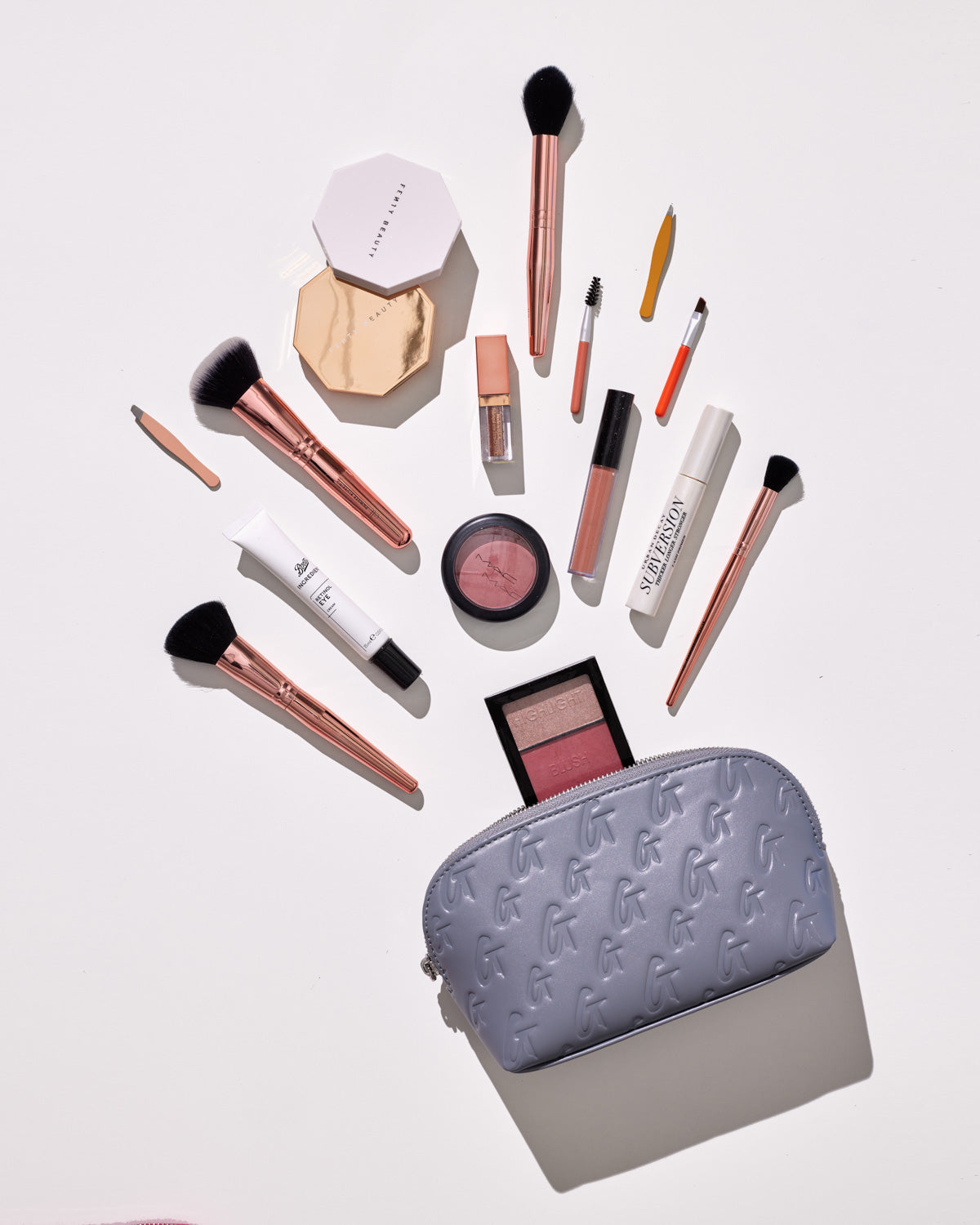 Gray monogram cosmetic pouch with makeup brushes, blush, lipstick, and other beauty products spilling out on a white surface.
