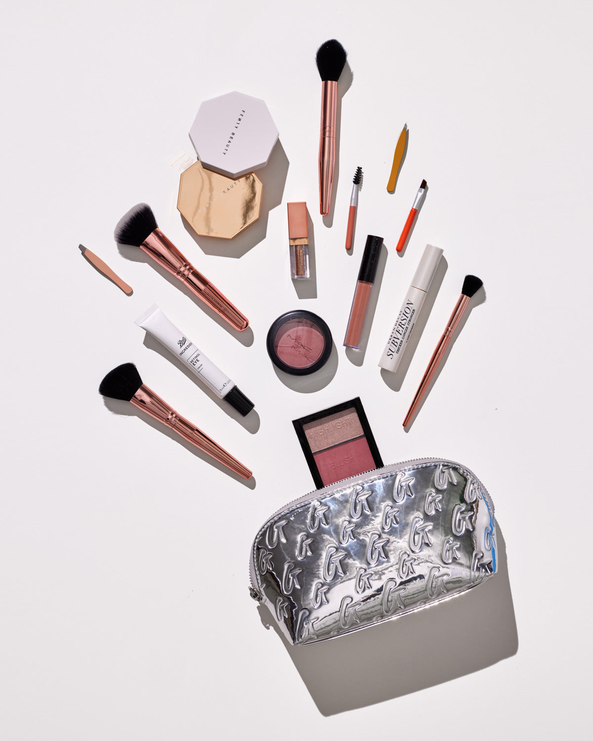 Silver metallic monogram cosmetic pouch with a repeating G pattern, open with makeup brushes, lip gloss, blush, and other beauty products spilling out.