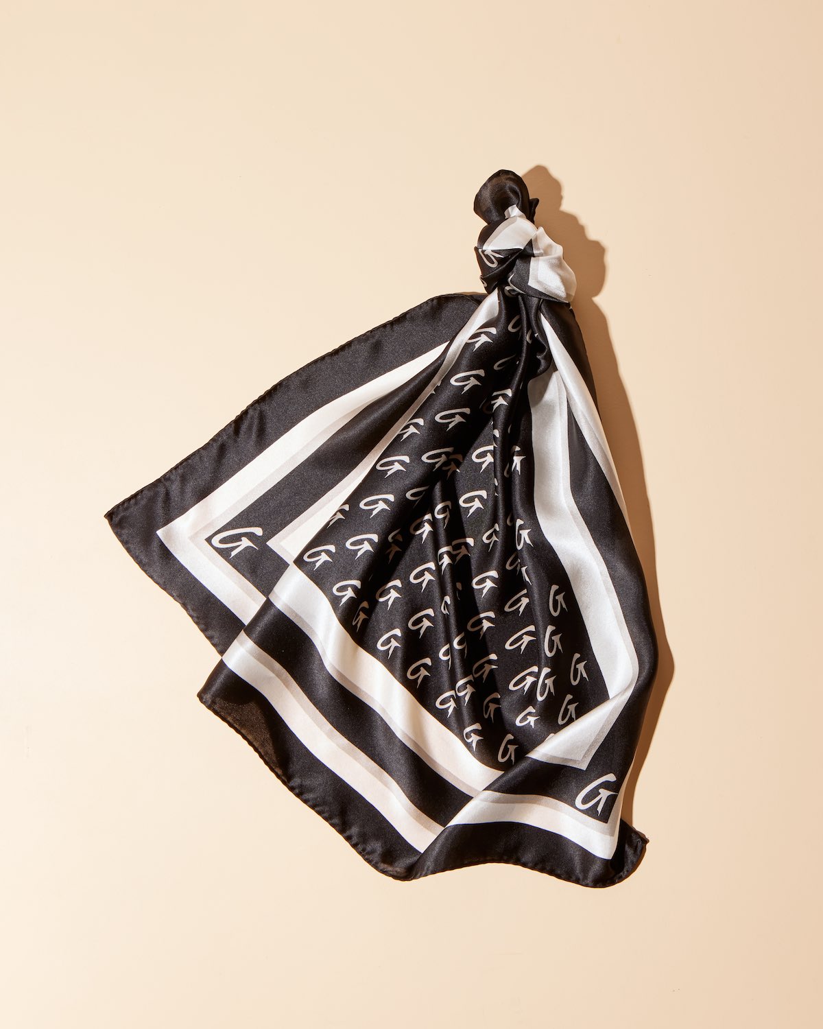 Black silk scarf with large monogram G pattern, bordered with white and black stripes, tied in a knot on a beige background.