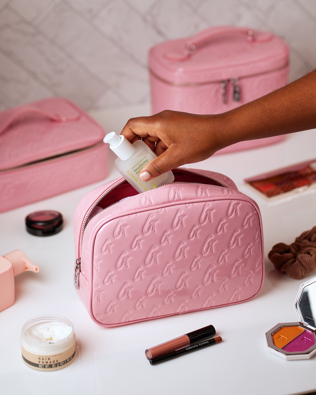 Pink monogram toiletry bag with embossed logo, open for skincare product, surrounded by makeup and accessories.