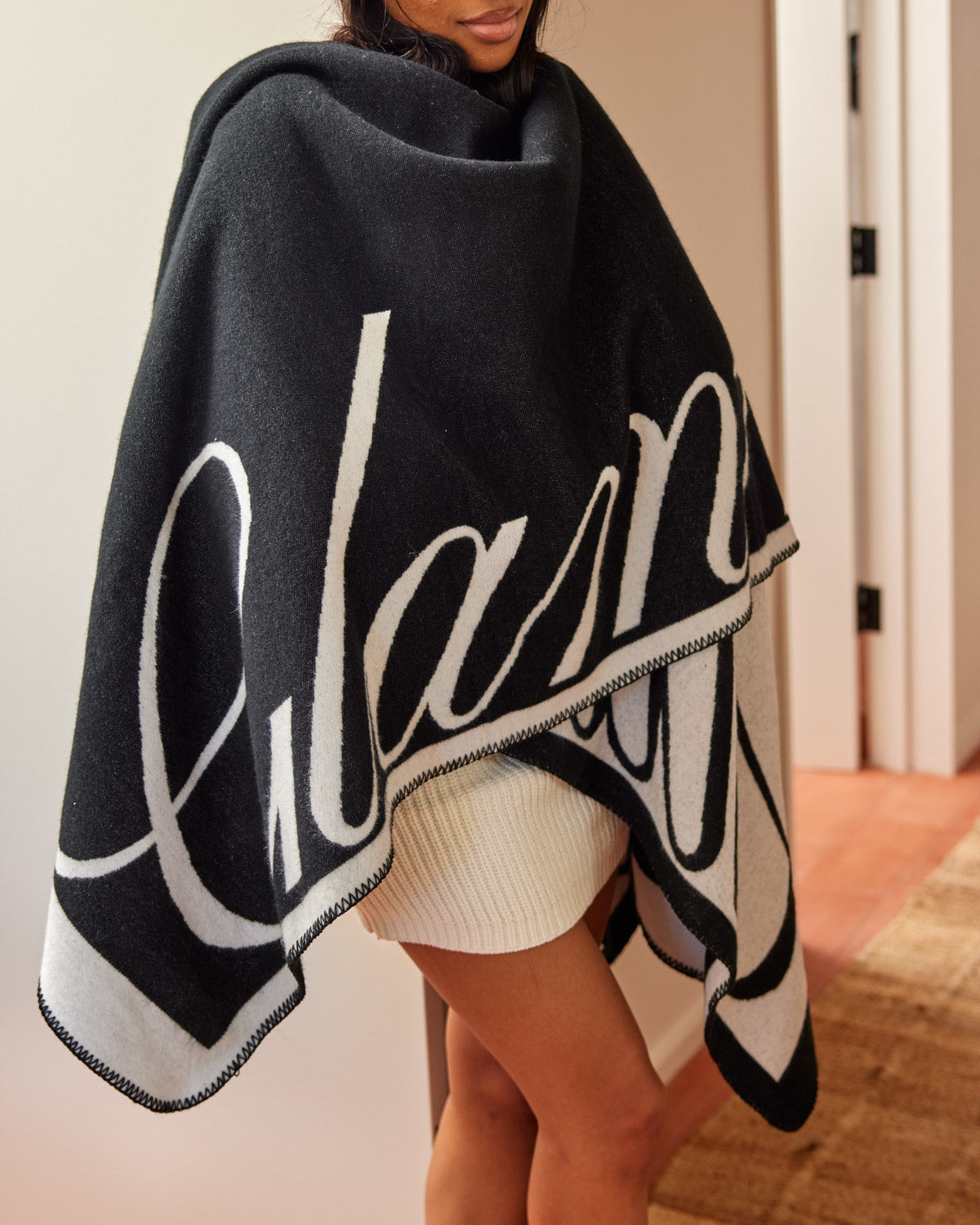 Black luxury throw blanket with white signature logo, draped over a woman's shoulders indoors.