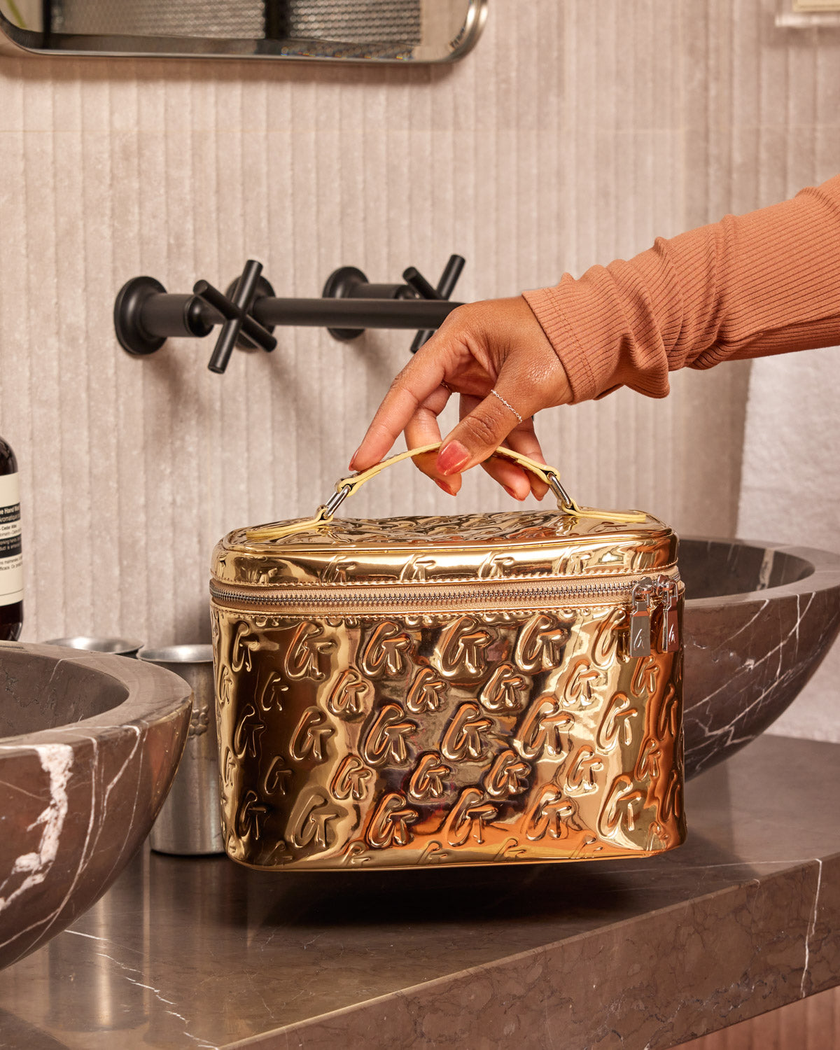 Large metallic gold monogram toiletry bag with embossed "G" pattern, held by a hand in a bathroom setting.