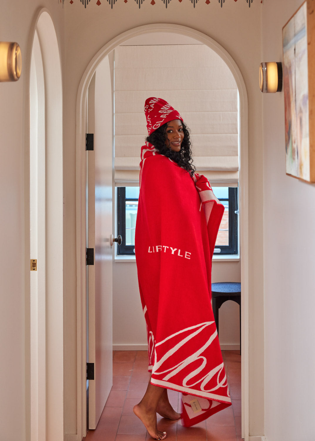 Woman wrapped in a red Signature Logo Luxe Throw Blanket and matching beanie, standing in a doorway with natural light.