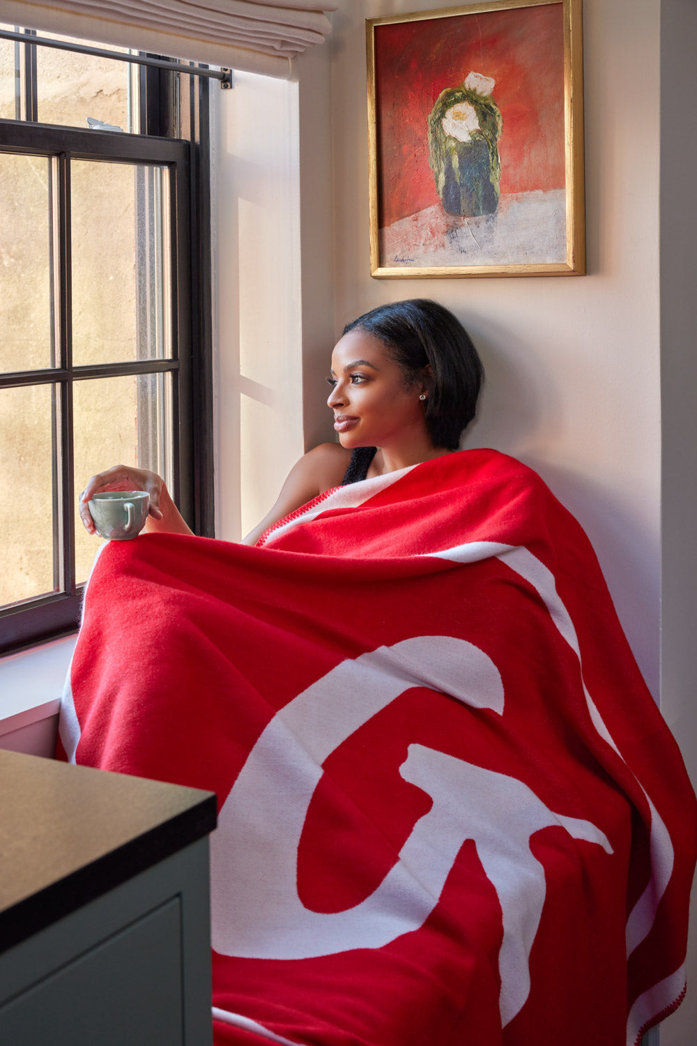A woman sitting by a window, wrapped in a red and white classic throw blanket, holding a mug, with a framed floral painting behind her.