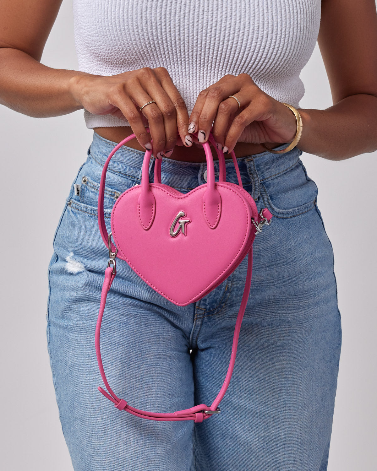 Pink heart-shaped mini bag with top handles, silver logo detail, and adjustable strap, held by a woman in a white top and jeans.