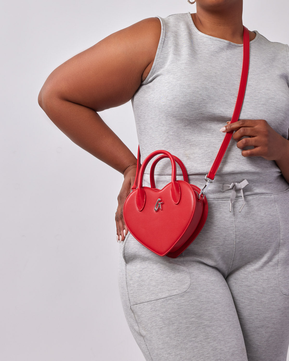 Red heart-shaped bag with a logo, worn crossbody style by a woman in gray athletic wear.
