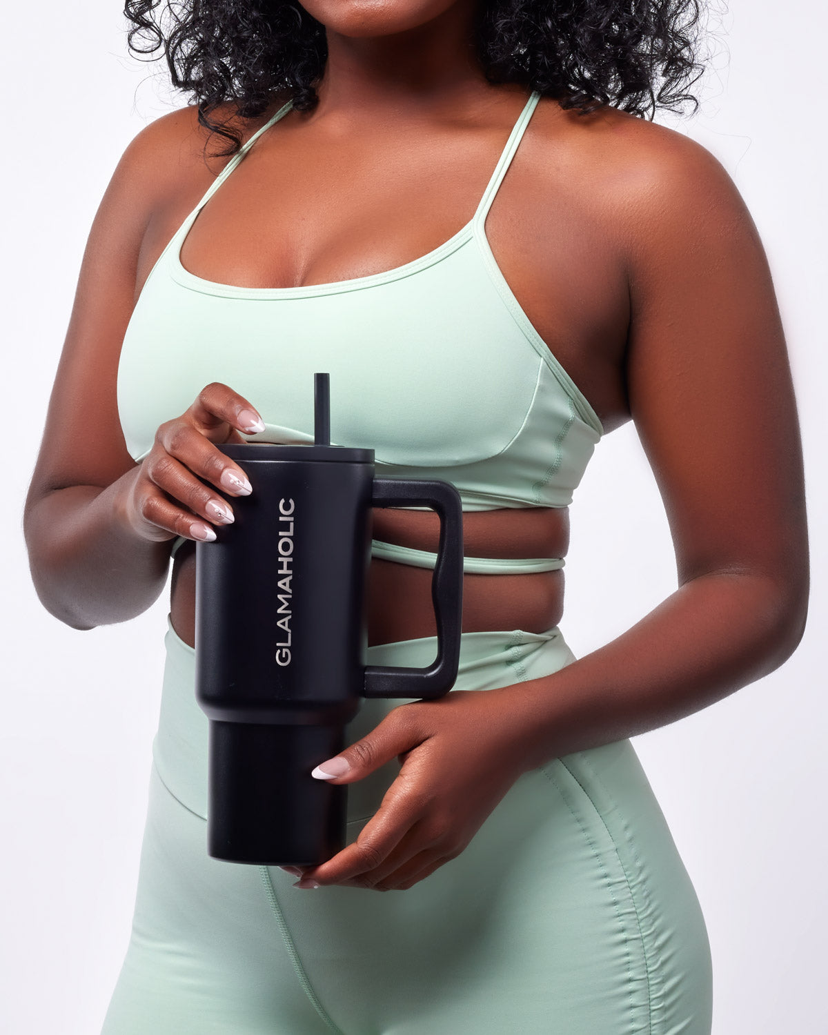 A woman in a mint green athletic outfit holds a black athletic tumbler with a straw, featuring the Glam-Aholic logo.