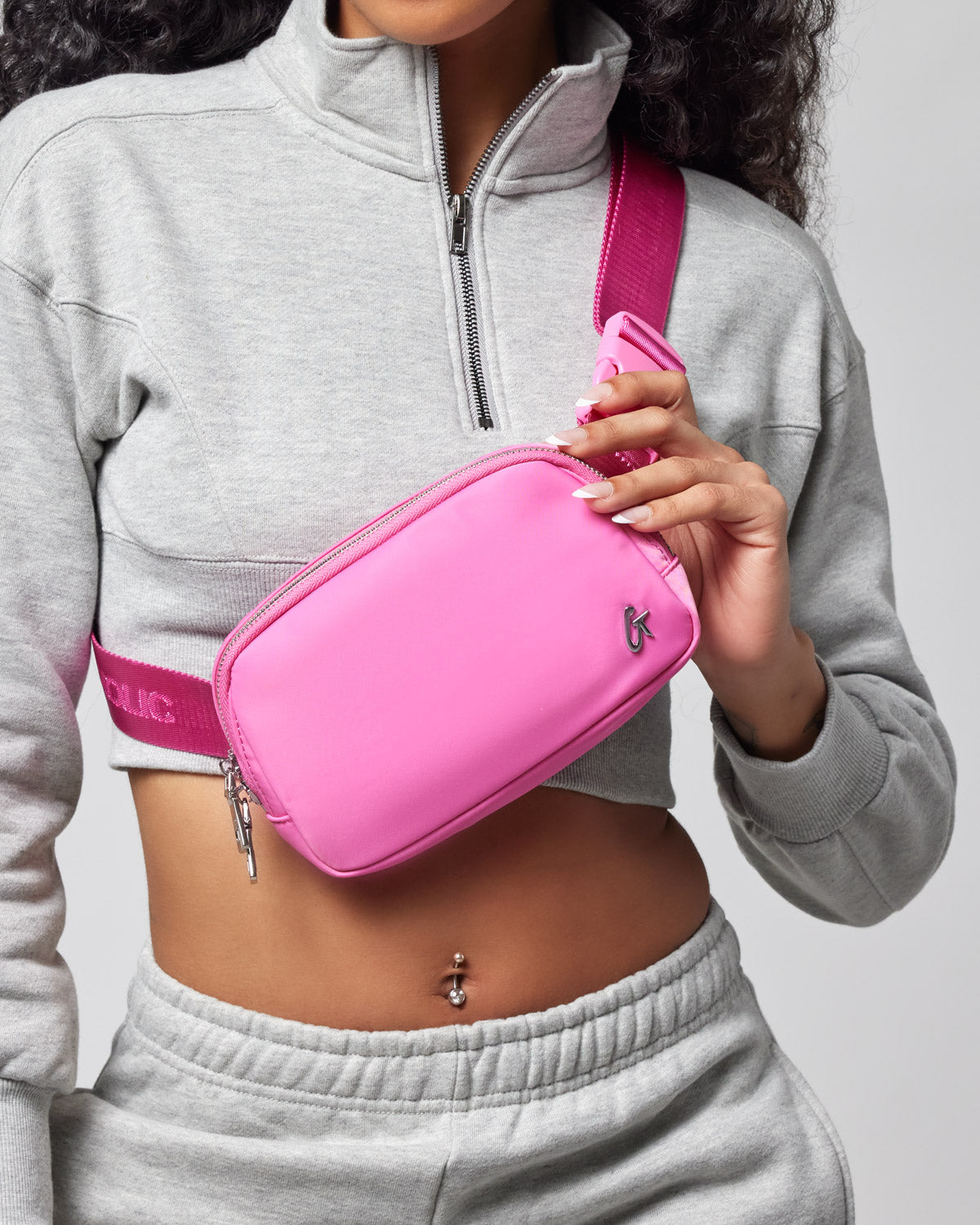 A woman wearing a gray cropped sweatshirt with a silver zipper, holding a bright pink nylon trio bag with a pink strap.