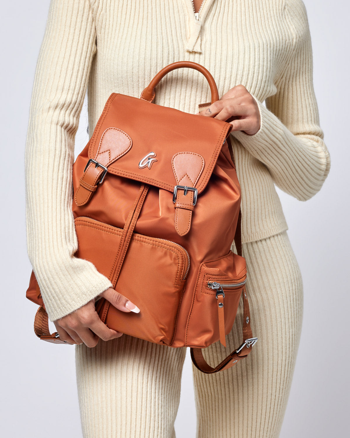 Brown nylon backpack with front zipper pocket, adjustable straps, and top handle, held by a person in a cream sweater.