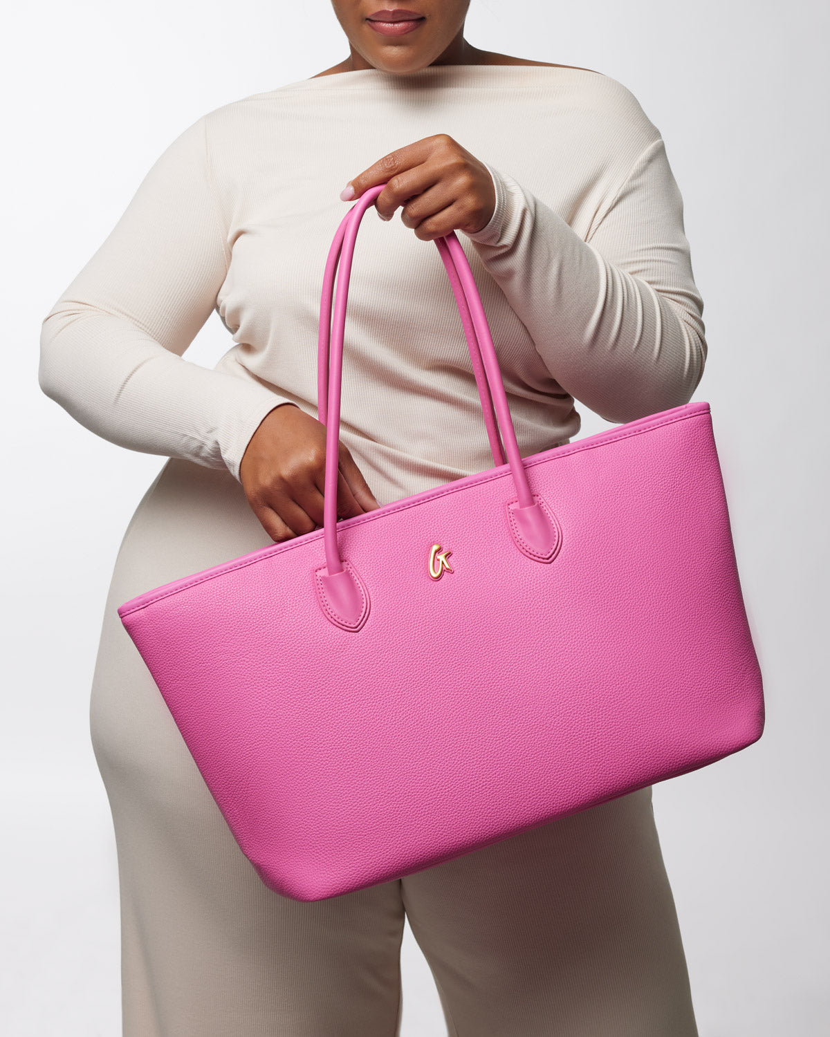Pink pebble-textured tote bag with dual handles and a small gold logo, held by a woman in a beige outfit.